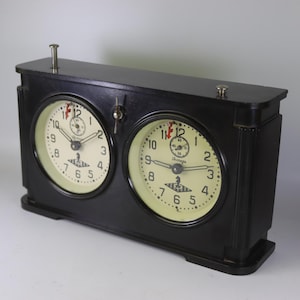 Könnte beinhalten: Schwarze Schachuhr mit zwei cremefarbenen Zifferblättern, die jeweils schwarze Ziffern und Zeiger anzeigen. Die Uhr hat einen Hebel und zwei Metallschieber oben.