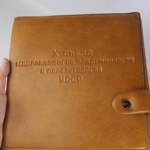 May include: A tan leather portfolio with embossed text. The text reads "X Съезд Микробиологов, Эпидемиологов и Паразитологов СССР". The portfolio has a button closure and is held by a hand.