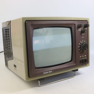 May include: A vintage Silelis 405 D portable television with a light beige plastic casing and a dark brown front bezel. The unit features a small screen, a large circular tuning dial, several smaller control knobs, and a metal wire stand base.