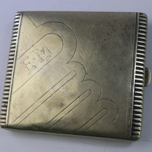 May include: A rectangular, silver-toned cigarette case with a decorative scalloped design and the initials "E.M." engraved on the front. The case has a ribbed edge and a clasp closure.