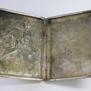 May include: Open silver-toned cigarette case with a detailed relief of a person and a horse. The case is rectangular with rounded corners and a clasp closure. The interior shows signs of age and wear.
