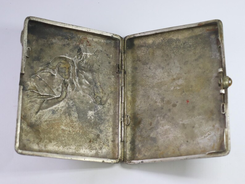 May include: Open silver-toned cigarette case with a detailed relief of a person and a horse. The case is rectangular with rounded corners and a clasp closure. The interior shows signs of age and wear.