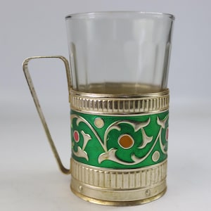 May include: A clear glass with a silver-colored handle and a decorative holder. The holder features a vibrant green enamel design with floral patterns and accents of red, orange, and white. The glass is approximately 4 inches tall.