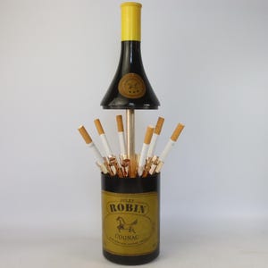 May include: A vintage Jules Robin Cognac bottle cigarette dispenser featuring a black cylindrical base with a gold label, a central metal rod, and a black and yellow bottle-shaped top cover that lifts to reveal multiple cigarette slots.