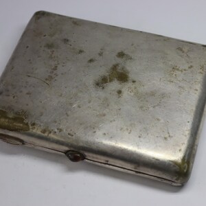 May include: A rectangular, silver-colored cigarette case with a tarnished, aged appearance. The case has rounded corners and a small, dark-colored clasp. The surface shows signs of wear and discoloration, with areas of green and brown oxidation.