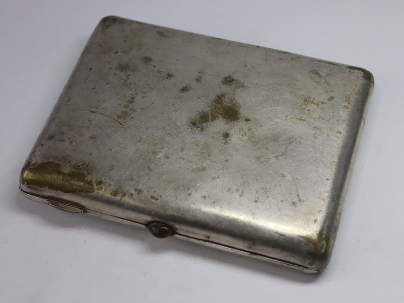 May include: A rectangular, silver-colored cigarette case with a tarnished, aged appearance. The case has rounded corners and a small, dark-colored clasp. The surface shows signs of wear and discoloration, with areas of green and brown oxidation.