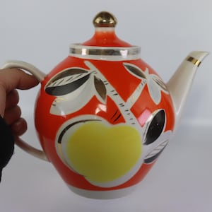 Könnte beinhalten: Eine Vintage-Teekanne mit einem leuchtend orangefarbenen Körper, verziert mit einem gelben Apfel und stilisierten Blattdesigns in Schwarz, Weiß und Gold. Die Teekanne hat einen weißen Ausguss und Griff sowie einen goldfarbenen Deckelknauf.