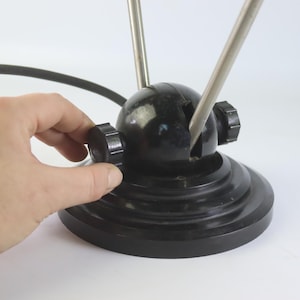 Puede incluir: Una antena de TV interior negra vintage con dos varillas cromadas ajustables. La antena tiene una base redonda y una esfera central con dos perillas. Una mano está girando una de las perillas. La antena está diseñada para recibir señales de televisión.