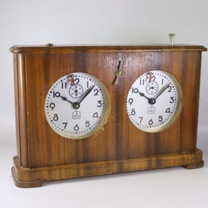 May include: A vintage wooden chess clock with two circular clock faces. The clock has a light brown finish and features black hands, white clock faces with black numerals, and a small metal lever on top. The clock is designed for timing chess games.