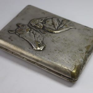 May include: A rectangular, silver-toned cigarette case with a detailed embossed design. The design features a horse's head and a reclining figure. The case has a tarnished appearance with gold-colored edges.