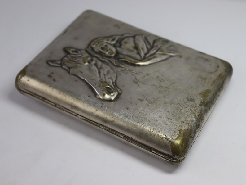 May include: A rectangular, silver-toned cigarette case with a detailed embossed design. The design features a horse's head and a reclining figure. The case has a tarnished appearance with gold-colored edges.