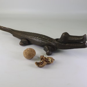 Può includere: Uno schiaccianoci a forma di alligatore in bronzo, con la bocca aperta, accanto a una noce intera e gusci rotti. Lo schiaccianoci misura circa 30 cm di lunghezza.