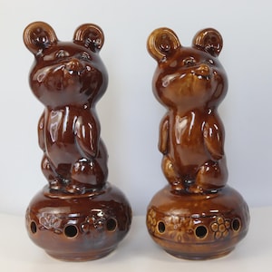 May include: Two glossy brown ceramic bear figurines standing on circular bases. Each bear has a smooth, reflective finish and rounded ears. The circular bases feature a series of small, evenly spaced circular cutouts and embossed floral patterns.