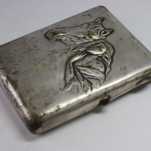 May include: A rectangular, silver-toned cigarette case with a detailed embossed design. The design features a horse's head and a woman's figure. The case shows signs of age and wear, with a tarnished appearance.