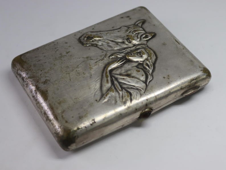 May include: A rectangular, silver-toned cigarette case with a detailed embossed design. The design features a horse's head and a woman's figure. The case shows signs of age and wear, with a tarnished appearance.