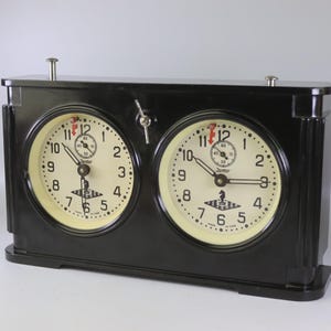 Könnte beinhalten: Schwarze Schachuhr mit zwei runden Zifferblättern, die jeweils ein cremefarbenes Zifferblatt mit schwarzen Ziffern und Zeigern anzeigen. Die Uhr hat einen Hebel und zwei Druckknöpfe. Auf dem Zifferblatt steht das Wort "Jantar".