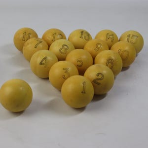 Puede incluir: Un conjunto de quince bolas de billar amarillas, cada una con un número negro del 1 al 15. Las bolas están dispuestas en un triángulo, con una bola separada. Las bolas tienen un acabado liso y mate.