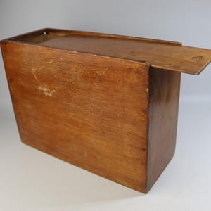May include: A rectangular wooden box with a hinged lid, stained in a warm brown colour. The box appears to be vintage, with visible wear and tear on the surface. The lid is partially open, revealing the interior of the box.