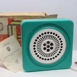 May include: A square teal plastic speaker with a white circular grille featuring a radial pattern of small holes. The speaker includes a white power cord with a plug, a cardboard box with red and yellow graphic details, and a paper instruction manual.