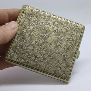 Op de afbeelding: Een vintage messing sigarettenetui met een sierlijk bloemmotief. De rechthoekige etui heeft een scharnierend deksel en een sluiting. Het oppervlak is bedekt met een herhalend ontwerp van bloemen en wervelende bladeren.