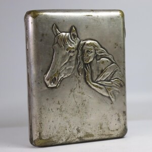May include: Vintage silver-toned cigarette case with a detailed relief of a horse's head and a woman's face. The case has a rectangular shape with rounded edges and a tarnished finish, showing signs of age.
