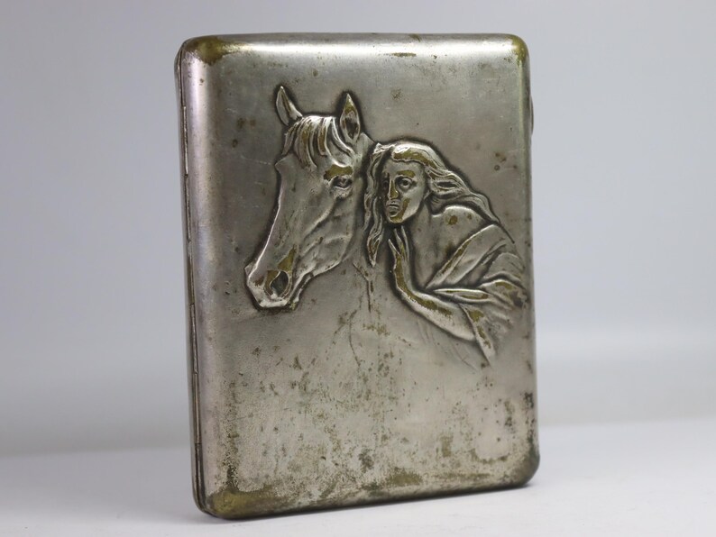May include: Vintage silver-toned cigarette case with a detailed relief of a horse's head and a woman's face. The case has a rectangular shape with rounded edges and a tarnished finish, showing signs of age.