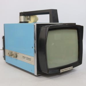 May include: A vintage portable television with a light blue metal casing, a black plastic handle, and a black screen frame. The front features the text "ЭЛЕКТРОНИКА ВЛ-100" and the side includes two silver control knobs with Cyrillic text labels.