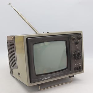 May include: A vintage portable television with a beige plastic casing and a black front panel. The device features a retractable silver metal antenna, a central screen, a large circular tuning knob, and several smaller control buttons. The text "SILOLIS 485 D-1" is printed in white on the lower front frame.
