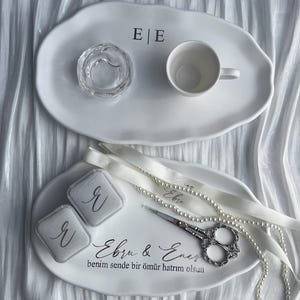Può includere: Due vassoi ovali in ceramica bianca. Il vassoio superiore presenta le lettere E|E. Il vassoio inferiore mostra il testo Ebru & Enes e benim sende bir ömür hatırım olsun, con due scatole per anelli grigie, forbici argentate e una collana di perle.