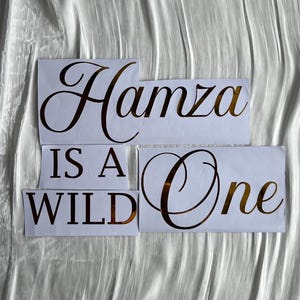 May include: White paper cutouts with gold lettering spelling "Hamza Is A Wild One" on a textured white background. The letters are in a stylish, elegant font, suitable for a birthday or celebration.