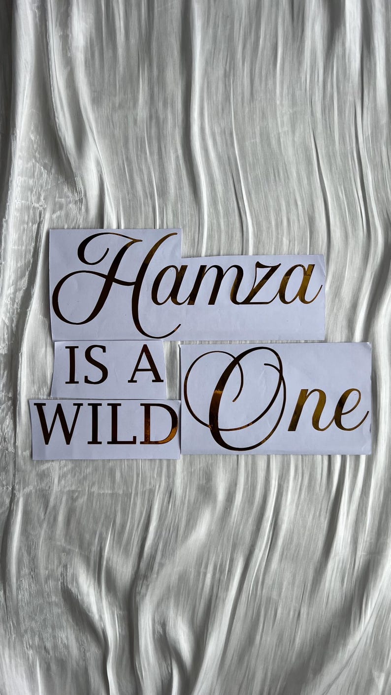 May include: White paper cutouts with gold lettering spelling "Hamza Is A Wild One" on a textured white background. The letters are in a stylish, elegant font, suitable for a birthday or celebration.
