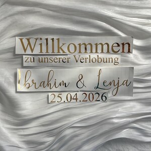 May include: Three rectangular signs with gold lettering on a white background. The top sign reads "Willkommen" and "zu unserer Verlobung" below it. The third sign says "Ibrahim & Lenja" and "25.04.2026".