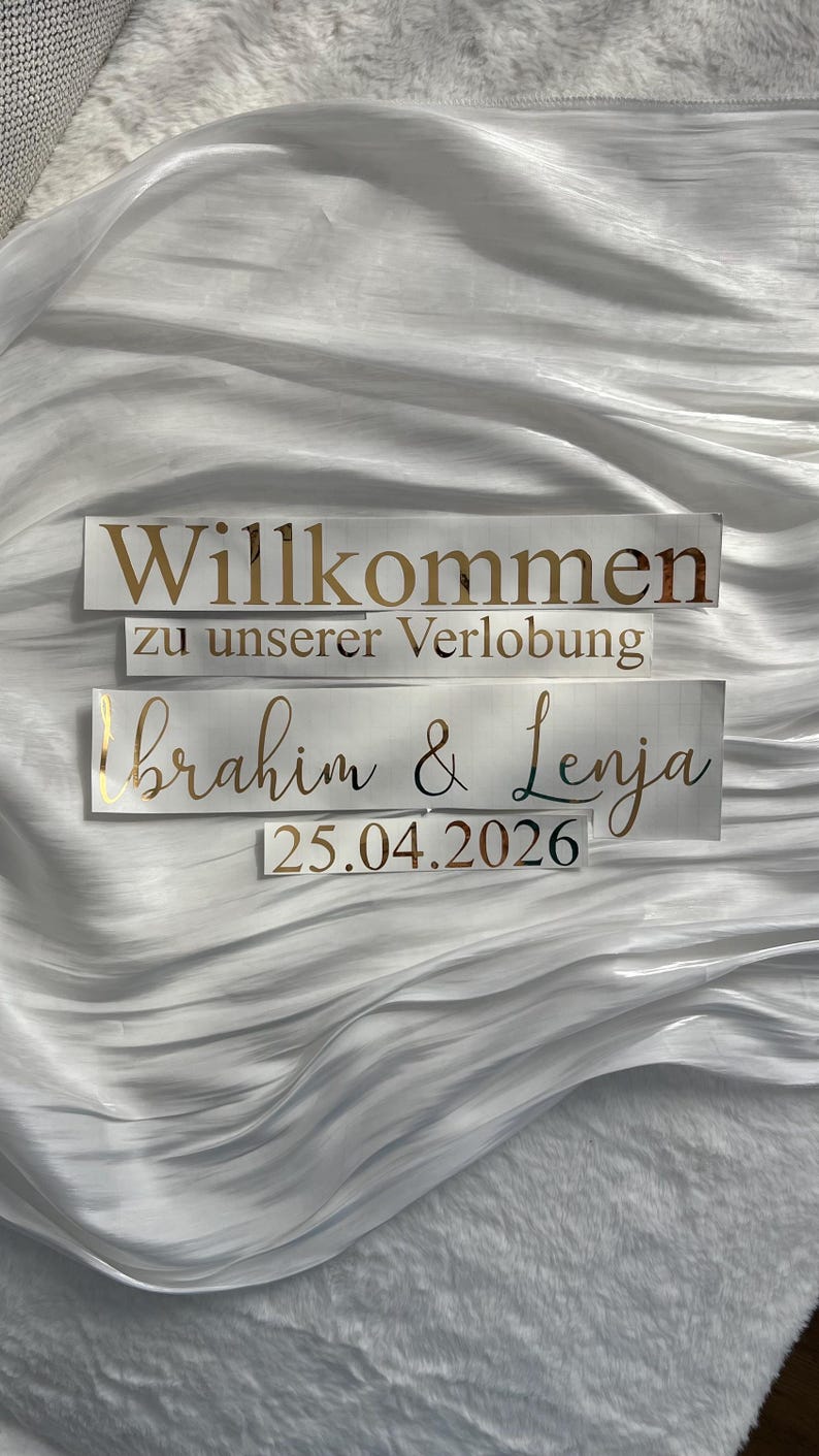 May include: Three rectangular signs with gold lettering on a white background. The top sign reads "Willkommen" and "zu unserer Verlobung" below it. The third sign says "Ibrahim & Lenja" and "25.04.2026".