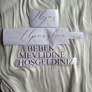 May include: White paper cutouts with gold lettering on a white, textured fabric. The cutouts read "Ilyas", "Alparslan'ın", and "BEBEK MEVLIDINE HOŞGELDINIZ". The fabric has a rippled, flowing appearance.