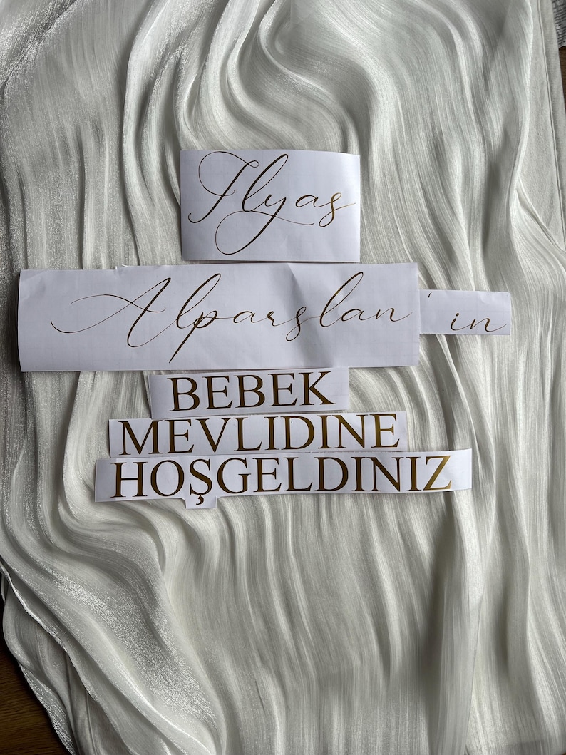 May include: White paper cutouts with gold lettering on a white, textured fabric. The cutouts read "Ilyas", "Alparslan'ın", and "BEBEK MEVLIDINE HOŞGELDINIZ". The fabric has a rippled, flowing appearance.