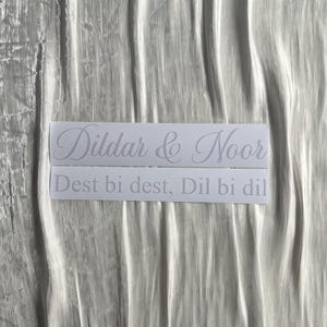 May include: Two white rectangular stickers with the names "Dildar & Noor" and the phrase "Dest bi dest, Dil bi dil" in a cursive font. The stickers are on a wrinkled, silvery-white fabric.