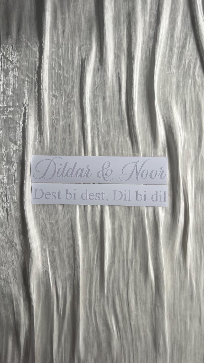 May include: Two white rectangular stickers with the names "Dildar & Noor" and the phrase "Dest bi dest, Dil bi dil" in a cursive font. The stickers are on a wrinkled, silvery-white fabric.