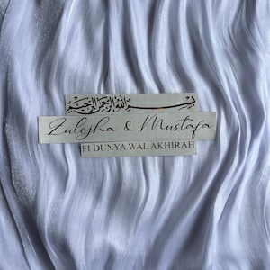 May include: White, flowing fabric with two rectangular stickers. The top sticker features Arabic script. The bottom sticker reads "Zulejha & Mustafa" and "FI DUNYA WAL AKHIRAH".