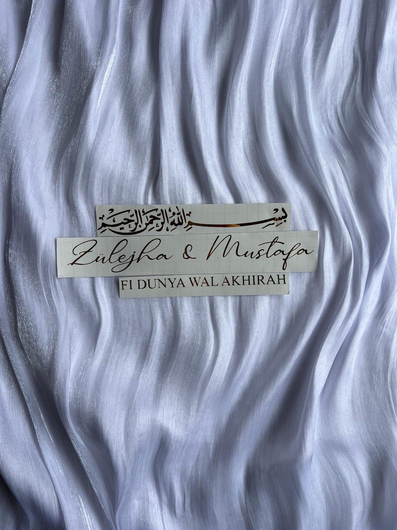 May include: White, flowing fabric with two rectangular stickers. The top sticker features Arabic script. The bottom sticker reads "Zulejha & Mustafa" and "FI DUNYA WAL AKHIRAH".