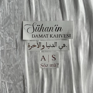 May include: A white background with three rectangular signs. The top sign reads "S&uuml;han'ın DAMAT KAHVESI" in black script. Below, Arabic text is displayed. The bottom sign has "A | S" and "S&ouml;z m&uuml;?" in black.