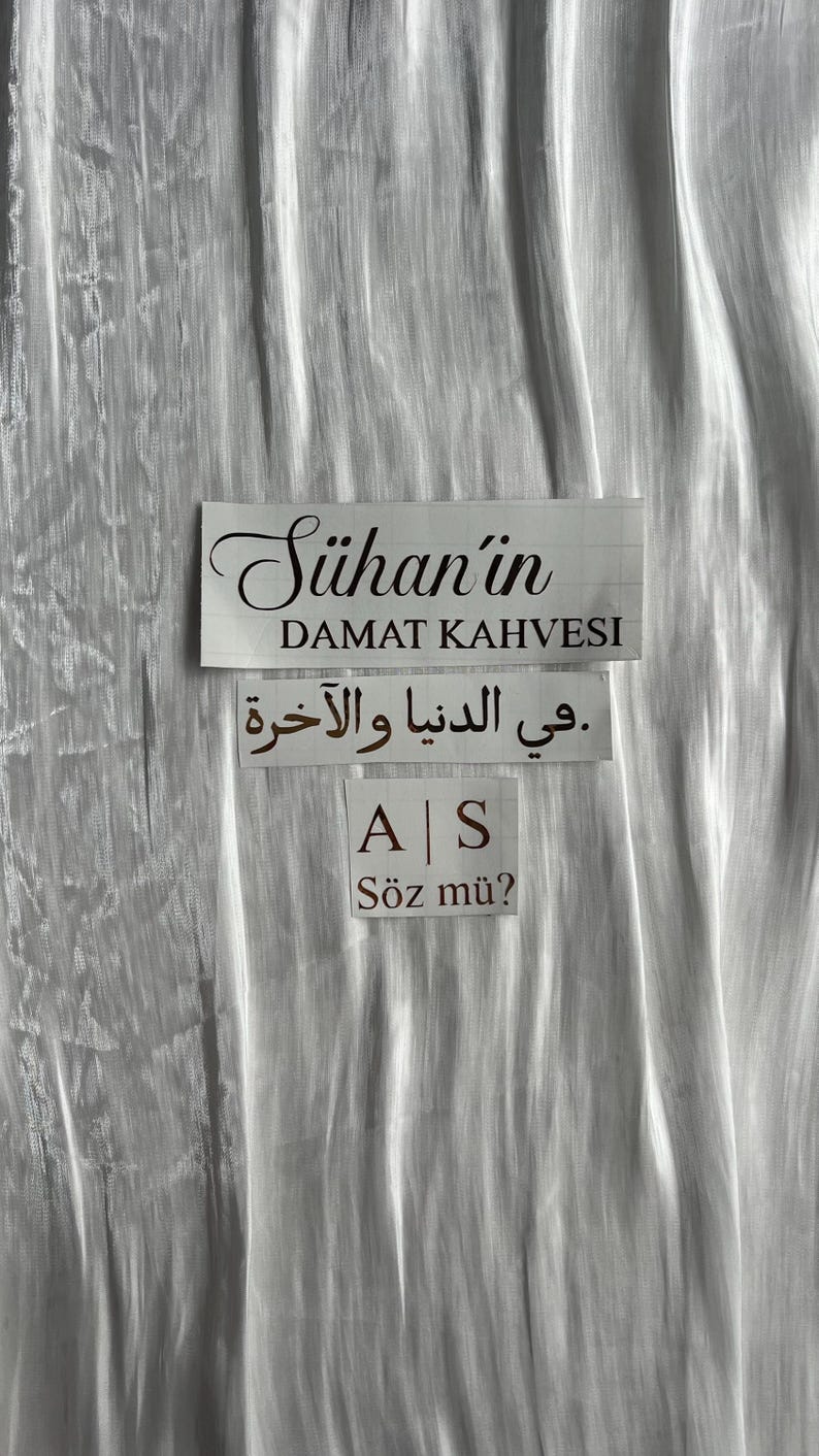 May include: A white background with three rectangular signs. The top sign reads "S&uuml;han'ın DAMAT KAHVESI" in black script. Below, Arabic text is displayed. The bottom sign has "A | S" and "S&ouml;z m&uuml;?" in black.
