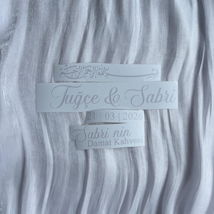 May include: White stickers with elegant script text on a textured white background. The stickers include names, a date, and the phrase "Sabri nin Damat Kahvesi". The overall theme is wedding or celebration.