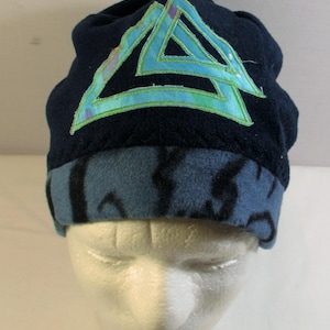 May include: A dark navy blue beanie featuring a large, light blue and green triangular geometric appliqué on the front. The brim of the hat is a contrasting light blue fabric with a dark, abstract, wavy black pattern printed throughout.