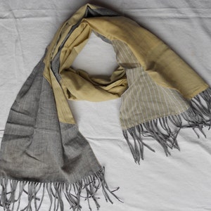 May include: A two-toned scarf with a gray and yellow color scheme. The scarf has a rectangular shape with fringed edges. One side features a solid gray fabric, while the other side is yellow with white vertical stripes. The scarf is draped on a white surface.