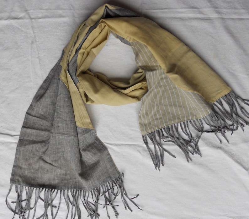 May include: A two-toned scarf with a gray and yellow color scheme. The scarf has a rectangular shape with fringed edges. One side features a solid gray fabric, while the other side is yellow with white vertical stripes. The scarf is draped on a white surface.