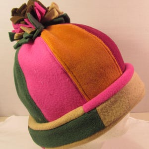 May include: A colorful fleece hat with a pom-pom. The hat features panels of pink, orange, green, and tan. The brim is tan with a pink trim. The pom-pom is made of green, pink, brown, and tan fleece.