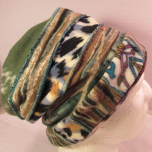May include: A multi-paneled beanie hat constructed from fleece fabric featuring various patterns including animal print, abstract stripes, and geometric shapes in shades of green, brown, blue, and cream, joined by visible teal overlock stitching.