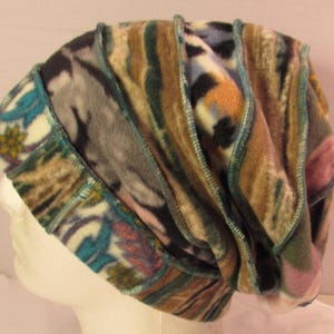 May include: A slouchy beanie hat constructed from multiple panels of fleece fabric in various patterns and colors, including brown, teal, cream, and black. The panels are joined with exposed teal serged seams, creating a textured, layered appearance.