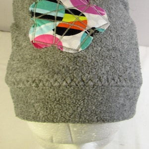 May include: A gray fleece beanie featuring a decorative fabric patch shaped like a flower. The patch displays a colorful pattern of circles in pink, teal, orange, and black, secured with visible zigzag stitching across the surface.