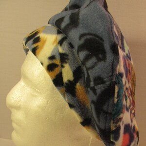 May include: A blue and multi-colored fleece beret with a patterned design. The beret is folded down and has a contrasting brown and black patterned fleece lining.